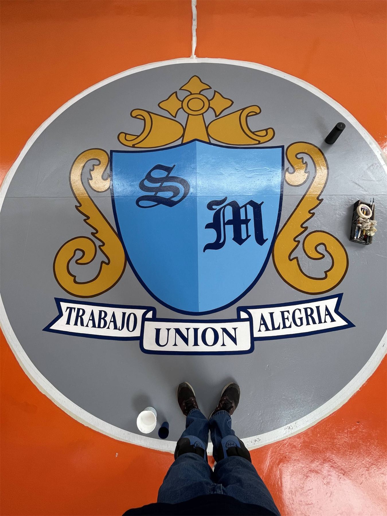 Escudo con el lema Trabajo, Unión, Alegría pintado sobre piso.