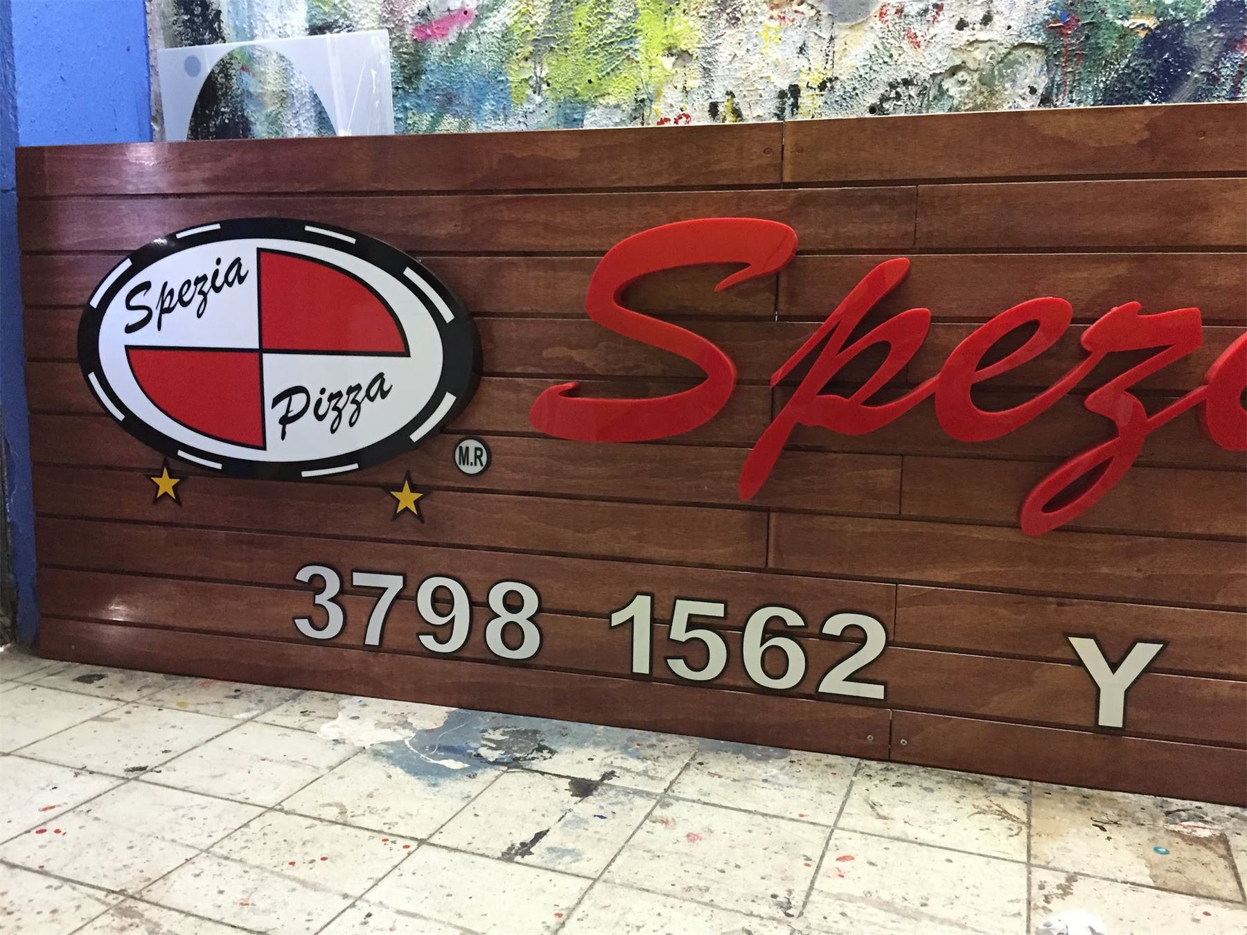 Letras volumétricas rojas de Spezia Pizza con logotipo sobre muro de madera.